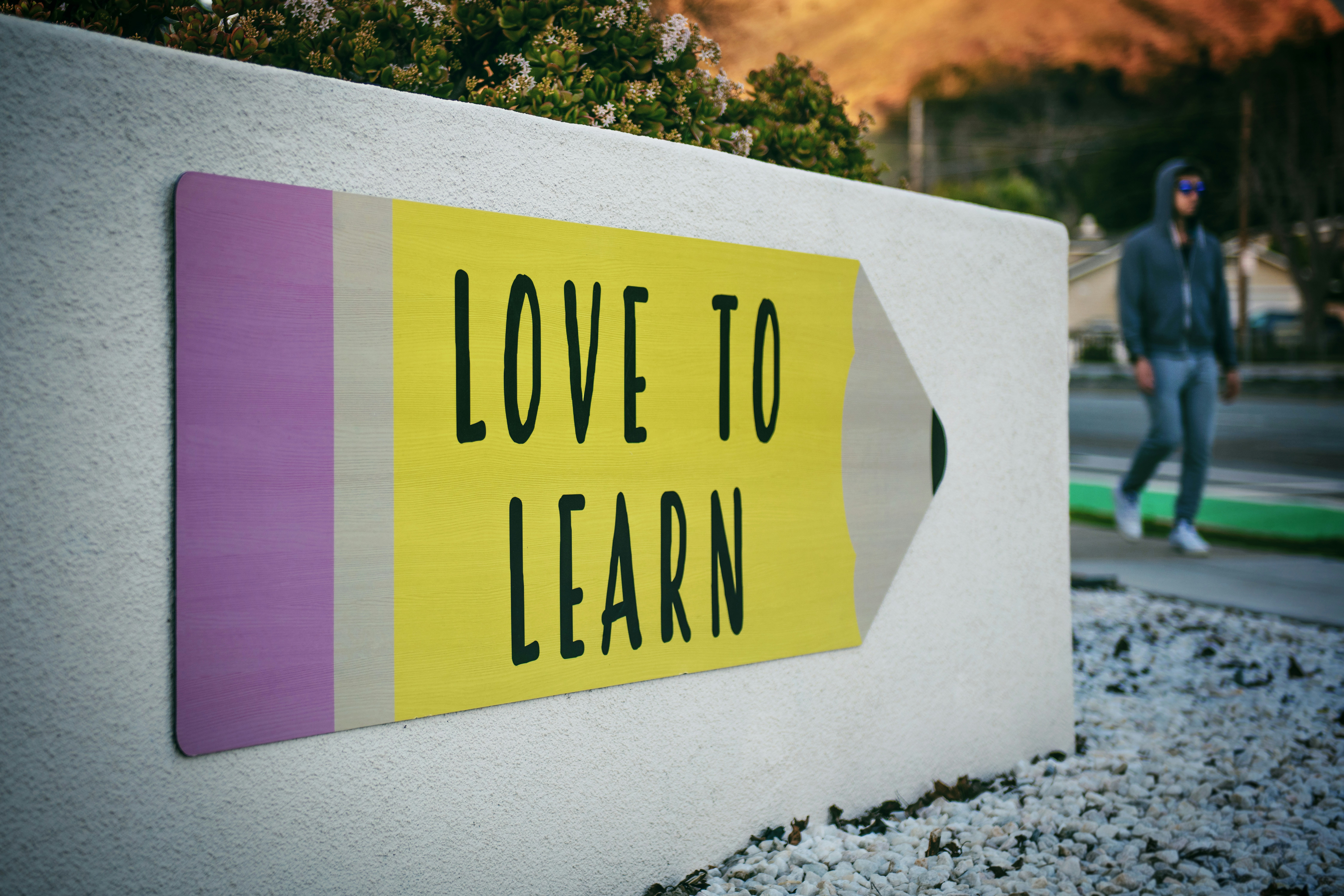 An outdoor sign on a concrete wall that reads love to learn with a man wearing a hoodie and shades walking by the sign