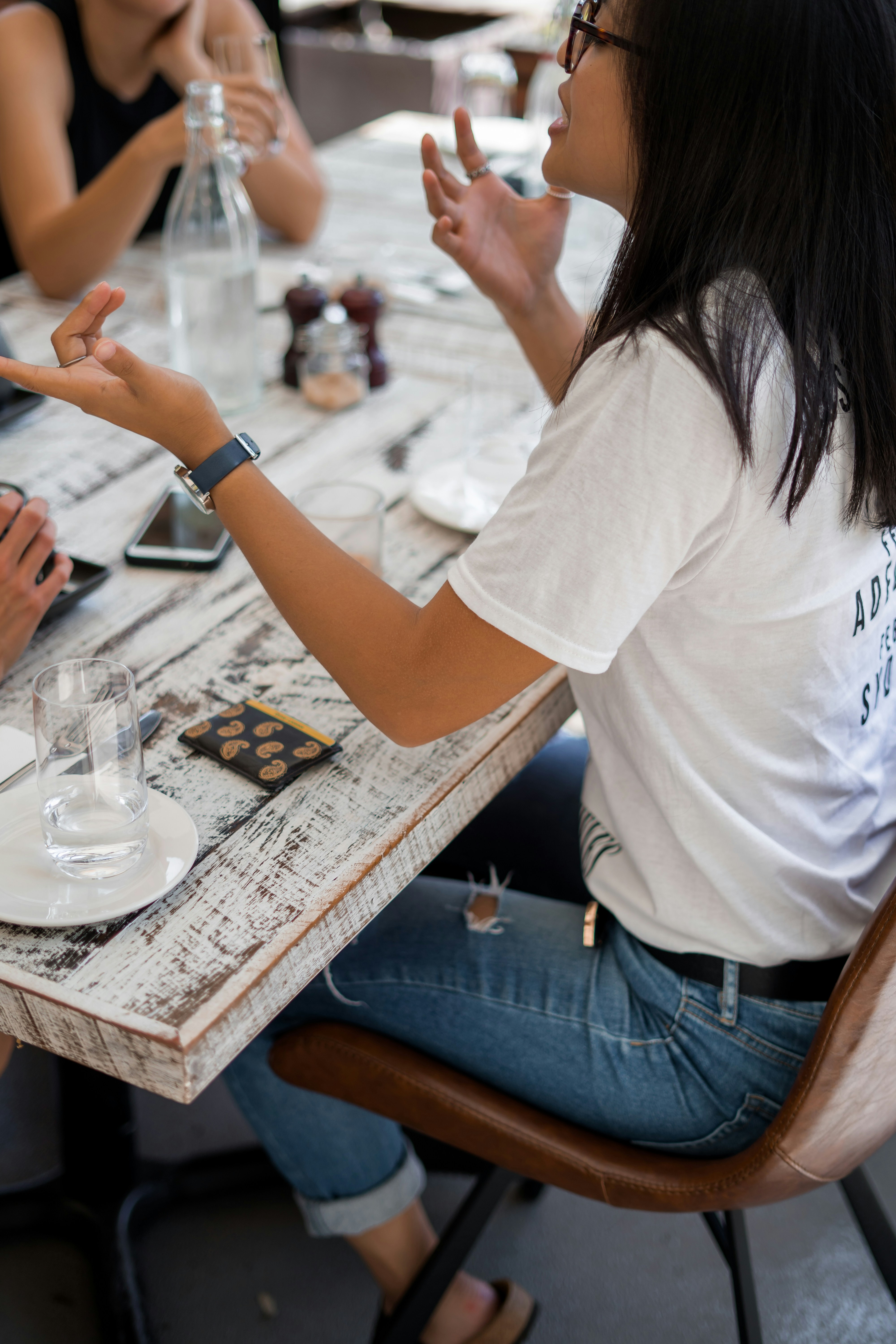 3 people sitting at a white wooden table outside talking. Person speaking is using their hands expressively