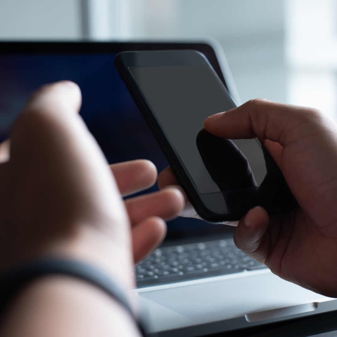 person holding a black cell phone with laptop on the table in front of them